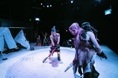 Cressida at the front right, back to us with Venus de Milon on his back, Achilles center, facing him, black mask and lacross stick, tribal women watching by a fire pit in the background and a pair of tents to the side. A video screen is in the upper right corner of the picture.