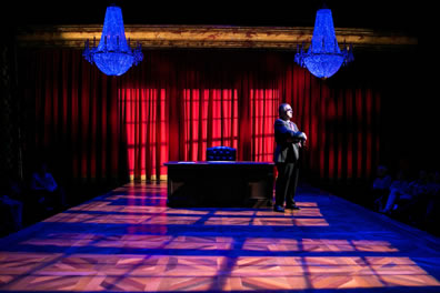 Scalia in a dark suit and tie stands at one end of his large oak desk with leather chair at the center underneath one of two chandeliers on a stage with the light coming through paned windows casting shadows on the parquet stage and red curtain in the background.