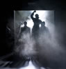 Coming through a large loading dock door with fog rolling in and a bight white spotlight from behind a woman in an overcoat, right fist raised, with other silhouette figures around her