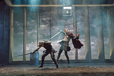 Production photo of Richard, with sword in hand, ducking out of the way of a sword swipe by Richmond, who is wearing an Elizabethan jerkin, skirt, and red cape.