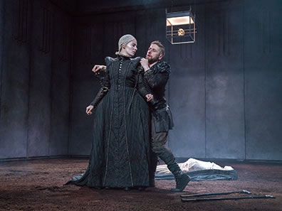 Production photo of Richard in black jacket and pants behind Lady Anne in simple black Elizabehtn dress. His hands are around his shoulder and his leg is crooked, the two crutches on the dirt bloor at his feet. Her head is turned looking at him. In the background is the white shrouded corpse of Henry VI.