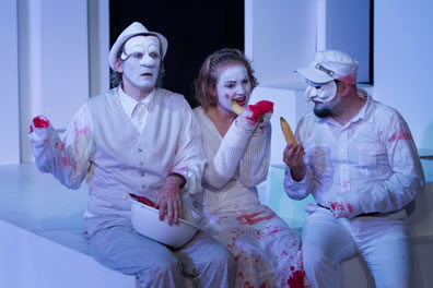 Marcus in fodor hat and vest and white face on the left holds a bowl; Lavinia in the center in white faced gnaws on an unpeeled banana; Titus in commedia mask and soldiers hat holds a banana. He and Marcus are missing a hand, Lavinia missing both hands, and blood stains are on their white clothes.