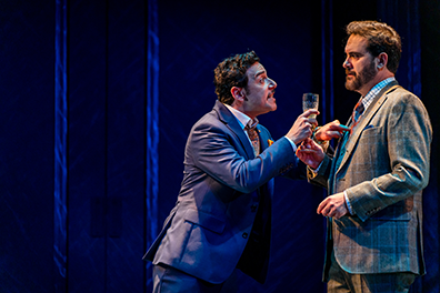 FOL-WInter's Tale Leontos with Camillo Leontes in blue suit and holding a champagne glass leans into and jabs his finger at the chest of the listening Camillo in gray three-piece suit.
