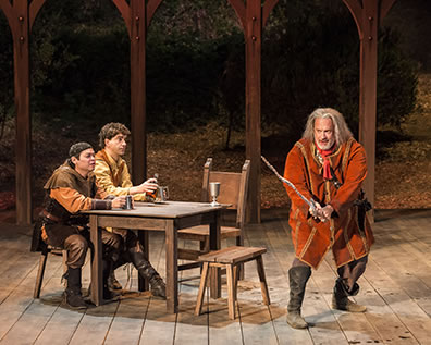 Production photo of Falstaff weilding a bent, hacked swoard as Poins and Hal sit at a table watching.