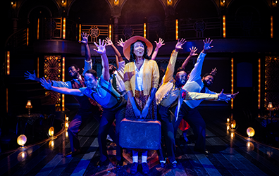 SIG-Play_On-Vy_on_A_Train Vy, wearing a jellow jacket, white blouse, orange patterend dress, white socks, plain shoes, and a wide brim hat stands with both hands holding a battered suticase in front of her as the ensemble behind her leans out with outstreched hands creating a starburst effect. Photo by Daniel Rader