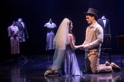 Maria and Tony kneel on the stage and holding hands, she in simple dress and wearing a wedding veil, he in brown work shirt and pants wearing a black tophat. In the background are maniquins with formal suits and nice dresses