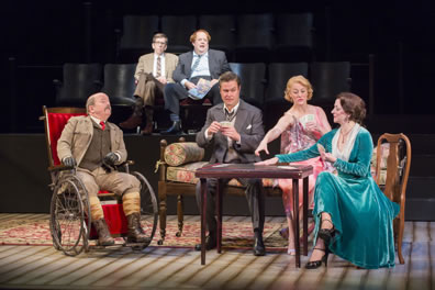 With Moon and Birdboot sitting in theater seats in the background, watching, Major Magnus dressed in brown hunters outfit, Simon Gascoyne in suit and vest, Felicity Cunningham in pink patterned flighty dress, and Cynthia Muldoon in richly turgouis dress, legs crossed, play cards at a table.
