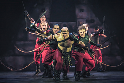 SYN-AntonyBoat Antony, torso bared and wearing red pants with an amrored belts and black shows, leans forward with ropes tied around his biceps, and angling out behind him are his crew, dressed in black breastplates, red pants, and black boots, all pulling ropes with one hand and holdin swords or poles int heir other hand. Their positions imply the bow of a boat. The pyramid set is in the shadowy background.